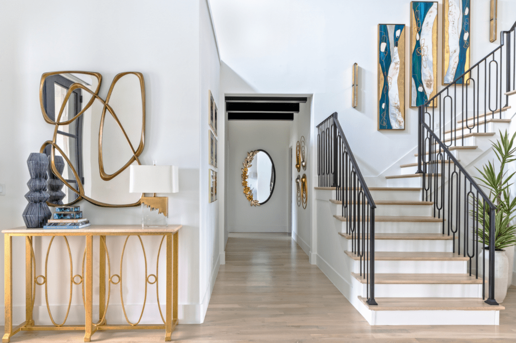 Modern entryway with gold accents, abstract art, light wood floors, and a black-railed staircase. Interior design by Baker Design Group.