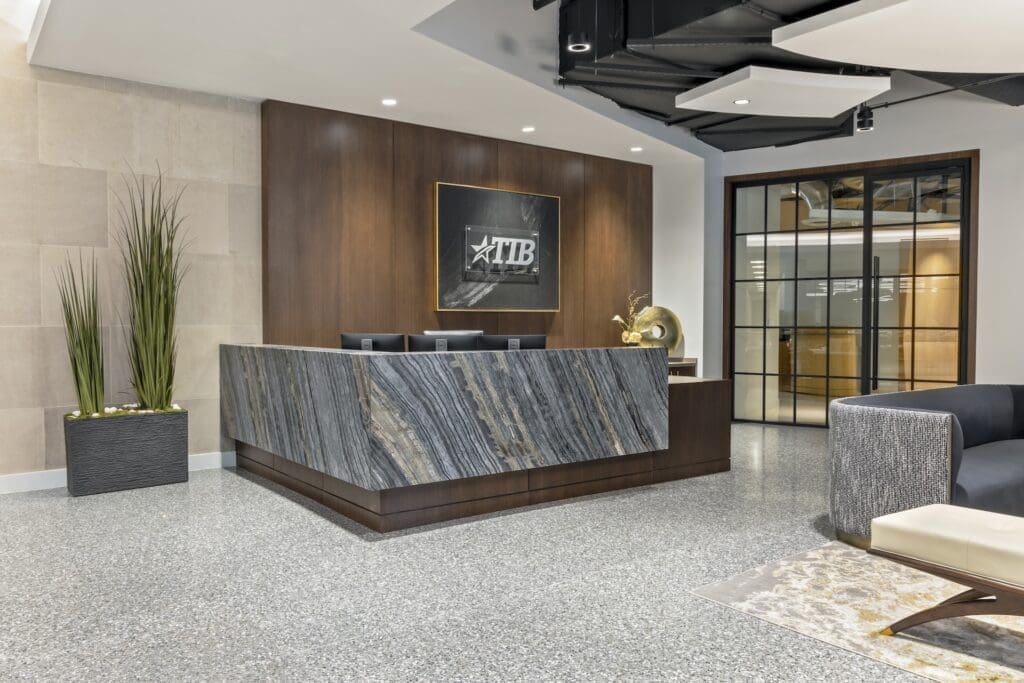 The reception lobby of a corporate bank featuring a sleek, dark stone reception desk, custom wood paneling on the walls, elegant circular ring chandeliers, and polished flooring designed by a premier commercial interior design firm.