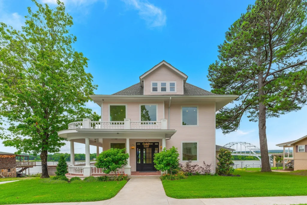 Exterior of a vibrant light pink 1920s home in Ozark, Arkansas, featuring a white front porch, second-story balcony, and riverfront views.