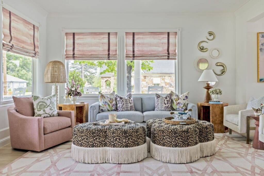 A vibrant living room interior design in an Ozark, Arkansas riverfront home, featuring a light blue sofa, pink armchairs, and custom leopard print ottomans with bullion fringe by Baker Design Group.