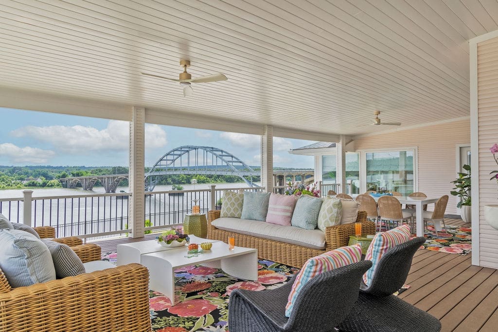A vibrant 1,600-square-foot wraparound porch design in an Ozark riverfront home, featuring weather-resistant rattan seating, colorful patterned cushions, and a stunning view of the river by Baker Design Group.