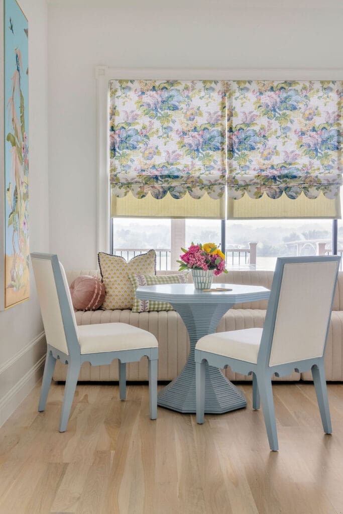 A vibrant kitchen nook interior design featuring a banquette-and-bistro setup with a blue pedestal table and custom layered scalloped fabric window shades by Baker Design Group.