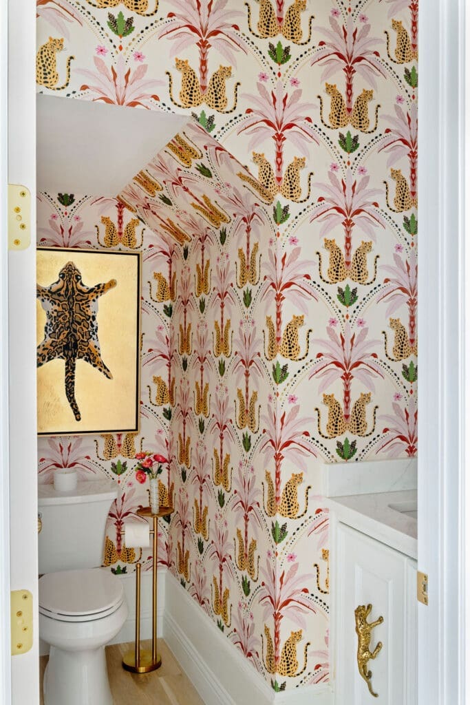 A bold, jewel-box powder bath interior design featuring pink and green tropical leopard wallpaper, gold accents, and framed animal art by Baker Design Group.