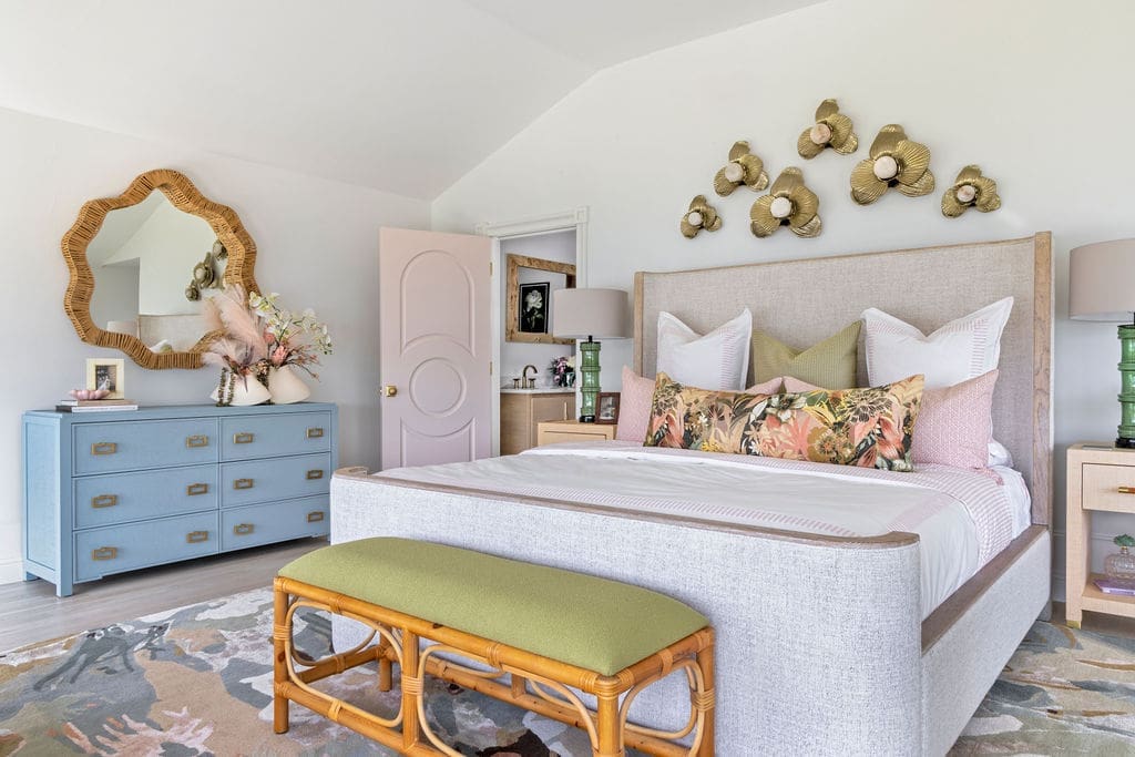 A vibrant, Miami-inspired owners' suite interior design in an Ozark, Arkansas home, featuring a light blue dresser, rattan accents, crisp white linens, and a green upholstered bench by Baker Design Group.